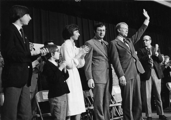 President Jimmy Carter waves to the audience who had come to hear him speak. Standing next to the President is Congressman David R. Obey and his family. Senator Gaylord Nelson can be seen applauding at the President's left.