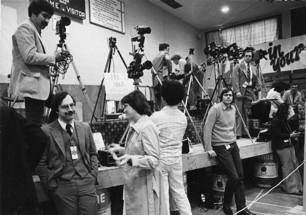 Press photographers set up their equipment in the press area before they cover a visit by President Jimmy Carter. A scoreboard hangs on the wall in the background.