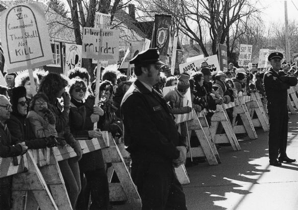 A group of pro-life demonstrators holding protest signs assembled to greet President Jimmy Carter during a visit to Wausau. The group is confined behind barricades and two police officers stand guard in the street. Trees and homes are in the background.