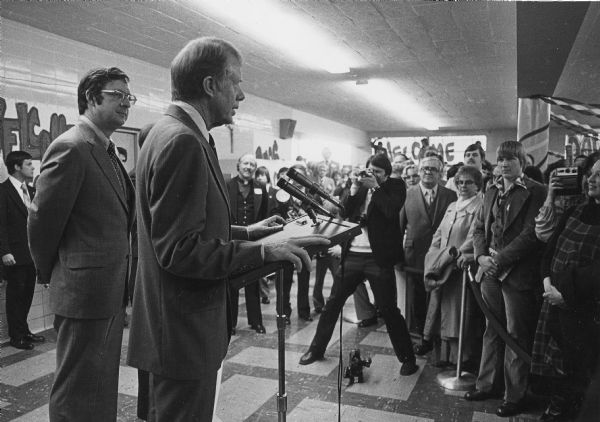 President Jimmy Carter speaks to a small group of well wishers while Congressman David R. Obey looks on. Photographers are in the crowd. The space is decorated with crepe paper and welcome signs.