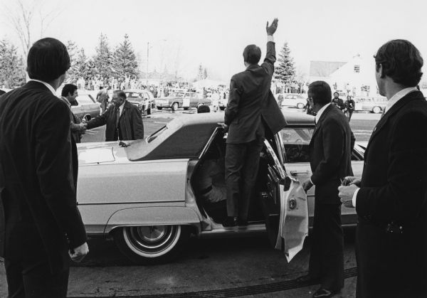 President Jimmy Carter stands in the open rear passenger door of a car to wave to a crowd of spectators during a visit to Congressman David R. Obey's hometown. Obey is shaking hands with a man on the left. A crowd of people stand near trees, a building, and automobiles in the background.