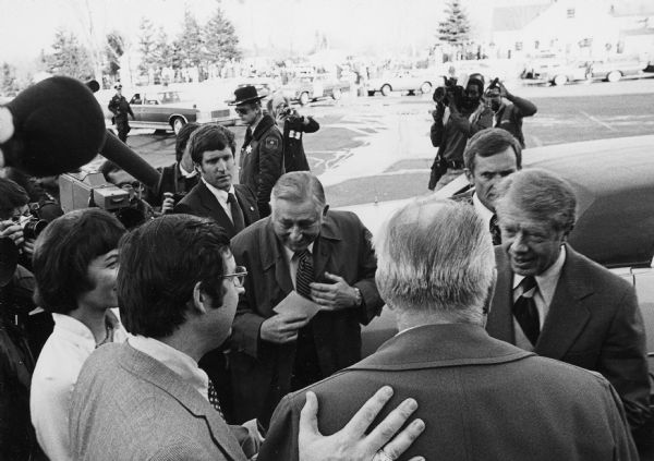 Congressman David Obey introduces a local resident (back to camera), to President Jimmy Carter. Many photographers stand around the edge of the group and a microphone is in the upper left corner foreground. Several police officers are on duty. A crowd of people stand near trees, a building, and automobiles in the background.