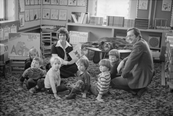 Wisconsin Congressman David R. Obey with pre-schoolers and their teacher at Ellis School, a program supported in part by Title I federal funding. Throughout his career Obey was a strong supporter of education. The children are in a semi-circle and were probably listening to the teacher reading a book. A bear skin with a stuffed head is on a table in the background.