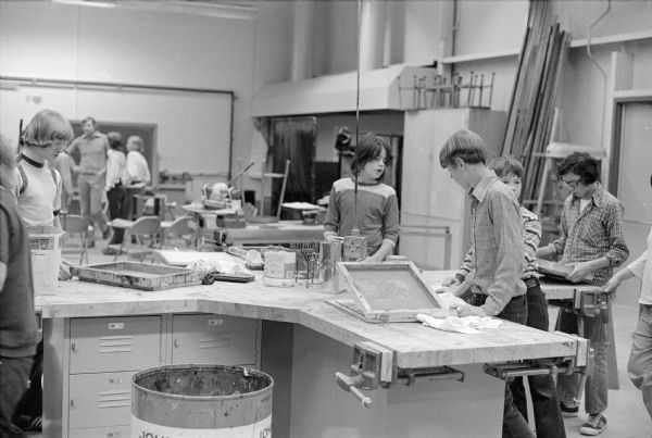 Vocational students at James Williams Junior High School work on a silkscreen project. The screens are laying on the work bench. More students, tables and chairs are in the background.
