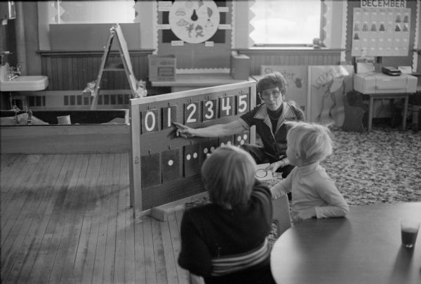 Teacher Dorothy Ledin teaching numbers to two pre-schoolers using a board with removable numbers and dots at Ellis School.