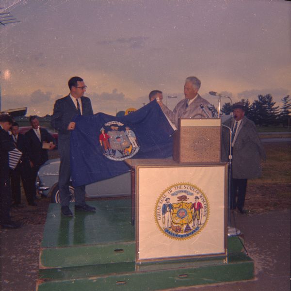 Assemblyman David R. Obey (left) and Governor Warren P. Knowles outdoors at an unidentified ceremonial event. The two men are holding the Wisconsin State Flag.