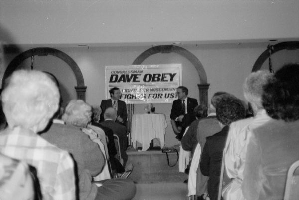 Wisconsin Congressman David R. Obey sits on stage with an unidentified guest speaker. There is a campaign banner behind them, and an audience is in the foreground. The event was probably a meeting of the Better Way Club in Wausau.