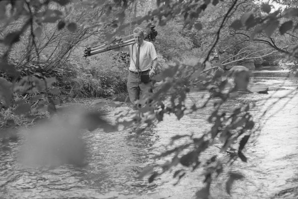 Photographer Fritz Albert wading through a stream in order to take a picture of congressional candidate David R. Obey meeting with a group of students near the stream. Although the location is unidentified it is likely near Wausau, Wisconsin.