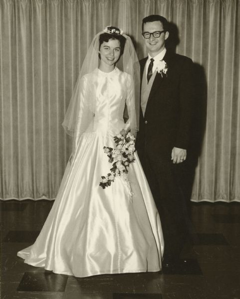 Joan and David Obey smile on their wedding day. She is wearing a wedding dress and he is wearing a suit with a vest.