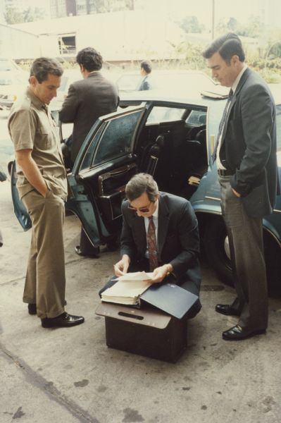 Congressman David R. Obey of Wisconsin crouches just outside of his car, reviewing the information in his briefing book during a congressional trip to Honduras.  Watching are Congressman Steve Solarz (left), and Congressmann Matt McHugh (right).