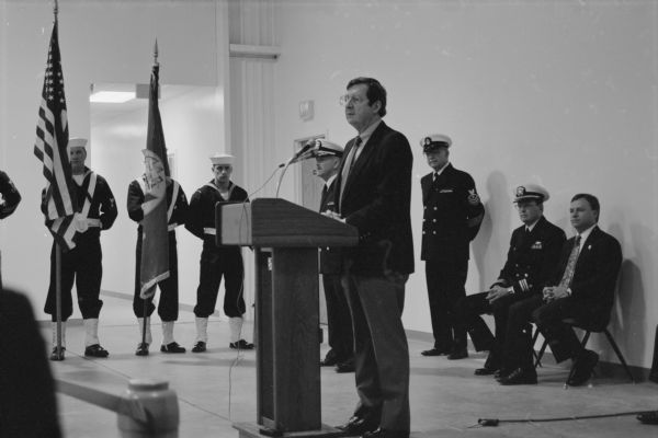 Congressman David R. Obey speaking at the podium at the dedication of the new Naval Reserve building. Three naval officers, a man in a suit, and three sailors are sitting or standing behind him. Two sailors hold flags.