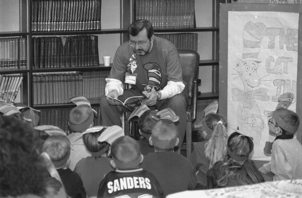 Congressman David R. Obey reads from "The Cat in the Hat" to young constituents who are wearing special hats. In the background are several shelves of books. On the right is a hand-drawn picture of the "Cat in the Hat."