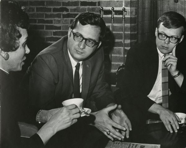 David Obey and Bill Bablitch listen as Mrs. Helen N. Van Prooyen speaks. They are drinking cups of coffee or tea. In the background is a brick fireplace, screen and utensils. The men are wearing suits.