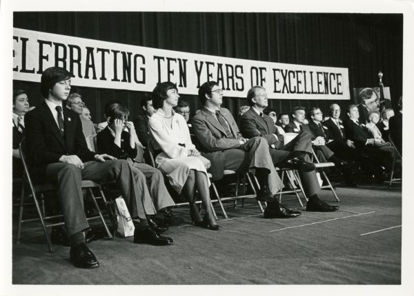 President Jimmy Carter listens to Senator Gaylord Nelson as he waits for his turn to speak. Sitting next to the President is Congressman David R. Obey and his family. A large banner in the background says, "Celebrating Ten Years Of Excellence." Next to it is a large portrait of Congressman Obey. Wisconsin dignitaries to the left of President Jimmy Carter who could be identified (all Democrats) are Governor Martin Schreiber, State Senator William A. Bablitch of Stevens Point, State Senator Fred Risser, and Congressman Alvin Baldus.
