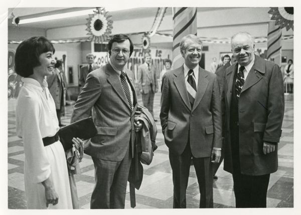 President Jimmy Carter poses with Congressman David R. Obey, his wife, Joan Obey and an unknown man. The space is decorated with crepe paper and welcome signs. Several people can be seen in the background.