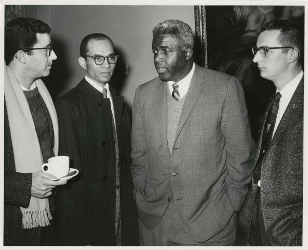 Stanley Zuckerman, Lloyd Barbee, Jackie Robinson, and David Obey are photographed as they are having a conversation. Zuckerman holds a cup, spoon and saucer. They are all wearing suits. Barbee is wearing a topcoat and Zuckerman, a scarf.