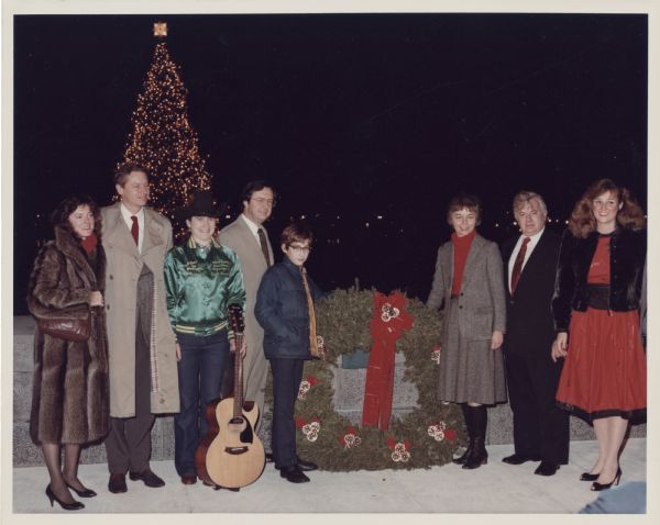 U.S. Representative David Obey, his wife Joan and their son Douglas pose with Connie G. Hicks (with guitar) and U.S. Representative Tom Petri. Between Douglas and Joan is a large wreath decorated with red bows and pine cones. Douglas flipped the switch to light the Capitol Christmas tree. Connie sang a Wisconsin version of "Oh Christmas Tree." The song "Christmas Spirit," a composition by Medford, Wisconsin's Bill Herrick was also sung by Nashville singer Julie Koblish, (she may be the woman on the right).