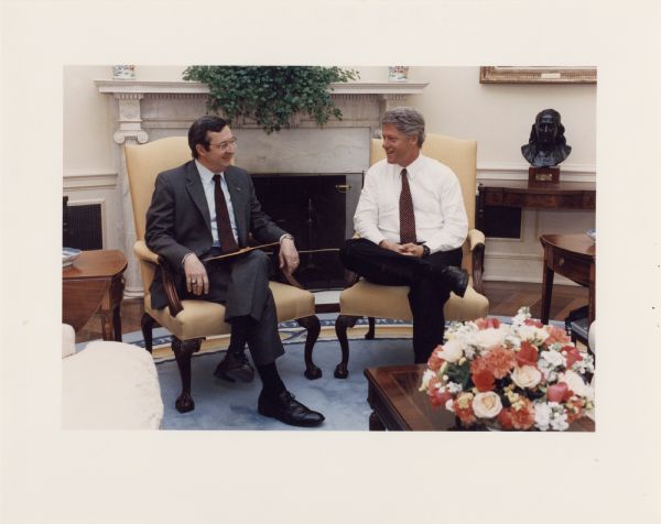 David Obey and President Clinton sit together in a meeting in the Oval Office. A fireplace is in the background.