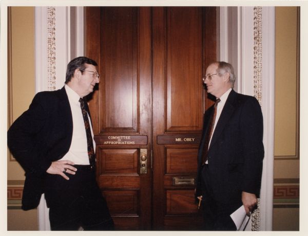 Congressman and Chairperson of the House Appropriations Committee David Obey and Scott Lilly stand on either side of the double doors leading into the office of the Committee on Appropriations. Lilly is holding papers and a pencil in his hands. He was a senior staff member of the House Appropriations Committee, executive director of the House Democratic Study Group, and chief of staff to Obey. Lilly also performed with Obey as part of the Capital Offenses, a bluegrass band formed by the congressman.
