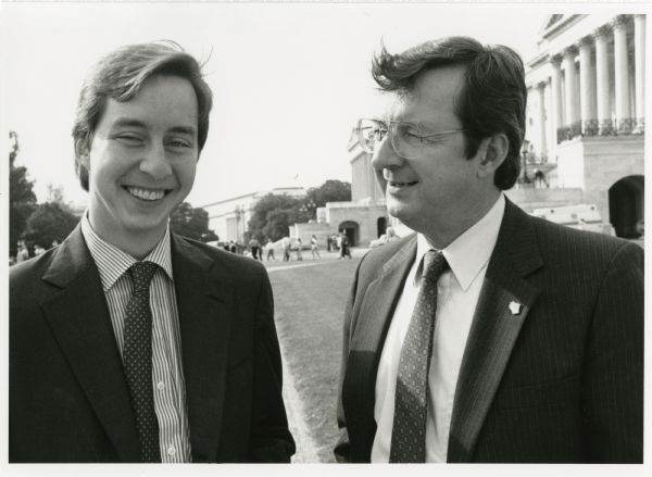 Congressman David Obey and his son Craig pose for a photo in the nation's Capitol. A line of people appear in the background.