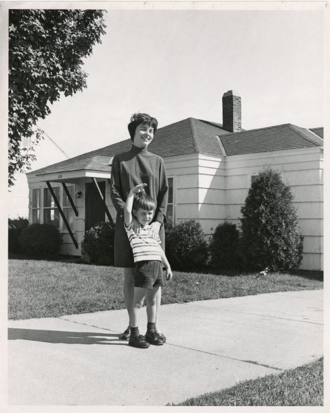 Joan and Craig Obey stand together in their driveway as Craig waves. The Obey's first house, at 515 North Ninth Avenue, and trees can be seen behind them.