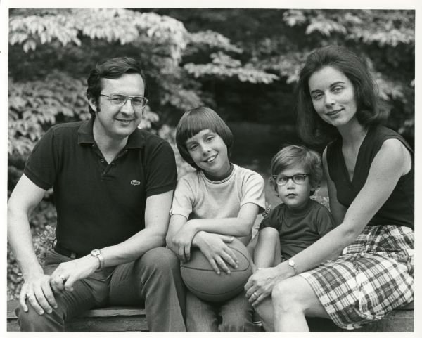 The Obey family poses for a casual outdoor portrait seated on a rock wall. (Left to right), David, Craig, Douglas and Joan. Craig is holding a basketball. Trees fill the background.