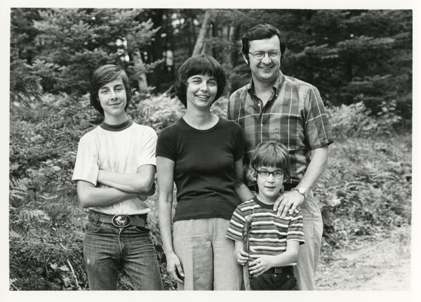 The Obey family poses, standing, for a casual outdoor portrait. (Left to right), Craig, Joan, Douglas and David. Douglas is holding a walking stick. Trees and foliage fill the background.