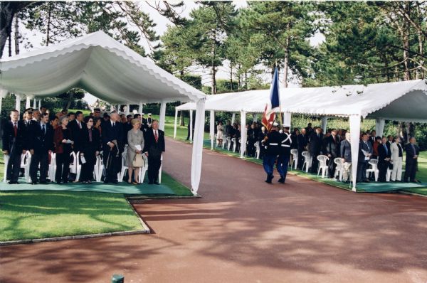 The Normandy Interpretive Center groundbreaking ceremony. Soldiers carrying the American flag march between two tents filled with guests. Participants in the groundbreaking include; Deputy Chief of Mission, U.S. Embassy France, Alejandro D. Wolff; U.S. Congressmen David L. Hobson and David R. Obey; ABMC Secretary MG John P. Herrling, USA (Ret); and U.S. Representative John P. Murtha. Joan Obey is standing to the right of her husband.