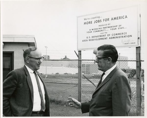 Wisconsin Democratic Party Chairman Louis Hanson and Wayne Lund[???] talk outside of a factory. The sign over Hanson's head reads, "We're creating...More Jobs For America. Produced by a Working Partnership of Your Community... Your State... and the U.S. Department of Commerce Area Redevelopment Administration. Lyndon B. Johnson." In the background is a chain link fence with barbed wire at the top and factory buildings.

