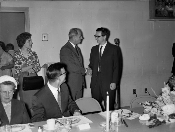 Congressman David Obey and Senator Gaylord Nelson shake hands as Joan Obey looks on. The occasion was a dinner, and a man and woman sit at a table with food, drink and candles in front of them. A flower arrangement is on the right. Behind Joan Obey is the door to the kitchen where two women are working.