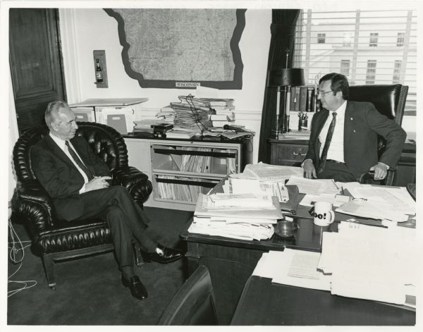 Prime Minister of Israel Shimon Peres and Congressman David Obey talk in Obey's office. Papers cover his desk and a large map of Wisconsin is mounted on the wall. A window behind Obey has a view of other buildings.