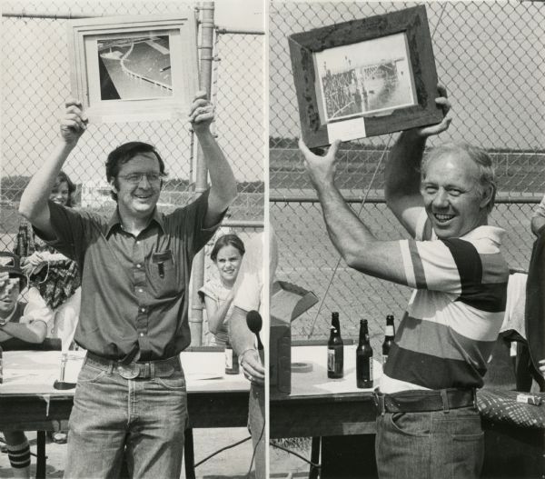 Before or after a Minnesota vs. Wisconsin Democrats softball game. Congressman David Obey holds up a concept rendering the future Richard I. Bong Memorial Bridge. Obeys' Minnesota counterpart holds up an old photograph of the bridge. In the background are tables, refreshments, people, a chain link fence and fields.
