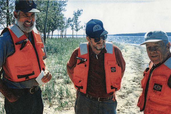 David Obey poses with Park Ranger Geoffrey Smith on the left and Martin Hanson on the right. They are standing on the beach with water and land in the background. Trees are on the left. All three men are wearing baseball caps and life preservers.