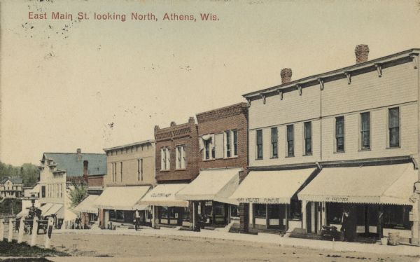 Colorized postcard view across street of the facades of furniture and clothing shops. Caption reads: "East Main Street, Looking North, Athens, Wis."