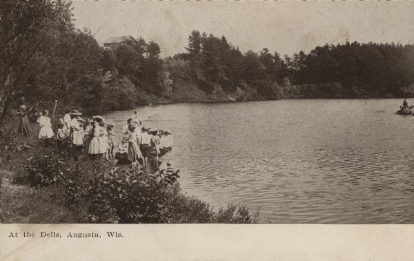 Photographic postcard view of a group of people gathered at the shoreline of a lake. One group is launching a boat near the shore. Another group is in a small boat out in the lake on the right. There is a building behind the trees on a hill in the background. Caption reads: "At the Dells, Augusta, Wis."