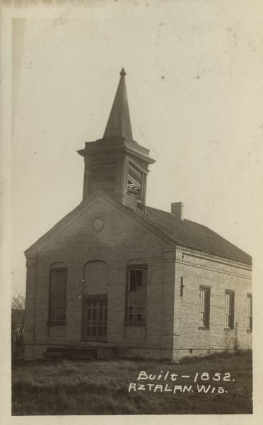 Photographic postcard of a three-quarter view of the exterior of an abandoned church. There are broken shutters in the belfry and a broken window in front. Caption reads: "Built — 1852, Aztalan, Wis."