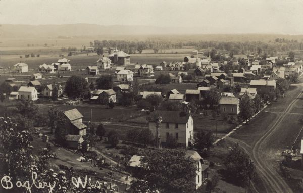 Elevated photographic postcard view of town showing homes and businesses. A windmill is in the foreground. The school is on the far edge of town on the left. There are unpaved roads and sidewalks throughout the town. Caption reads: "Bagley, Wis."