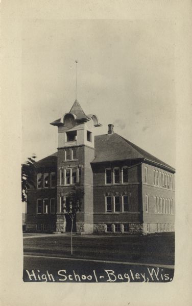 Photographic postcard of an exterior view of the high school, which has a bell tower above the entrance. There is a small tree in the schoolyard. Caption reads: "High School, Bagley, Wis."