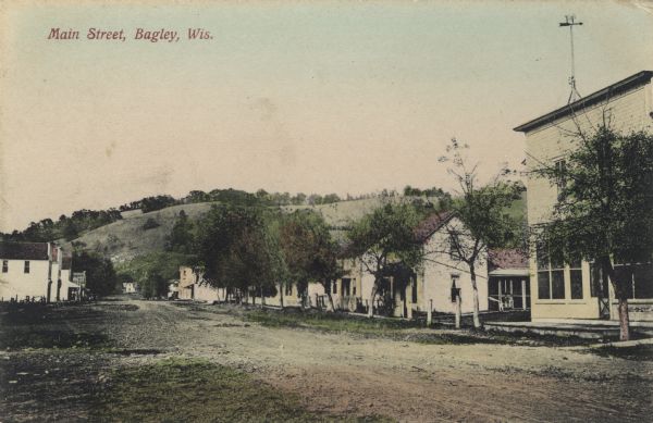 Colorized photographic view of the central business district. Homes, businesses and trees line the unpaved street, and there is a hill in the far background. Caption reads: "Main Street, Bagley, Wis."