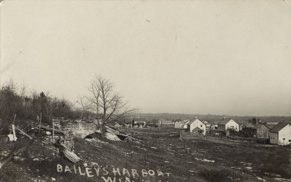 Photographic postcard view across rocky field of dwellings and other buildings along Lake Michigan. Along the left is a rocky ledge. The shoreline of the harbor is on the right. A tree-lined hill or ridge is in the far background. Caption reads: "Baileys Harbor, Wis."