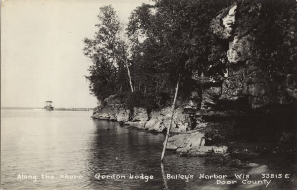 Photographic postcard view along the rocky shoreline of Lake Michigan towards a long pier that jutts out behind the rocky point. Caption reads: "Along the Shore, Gordon's Lodge, Baileys Harbor, Wis. Door County."