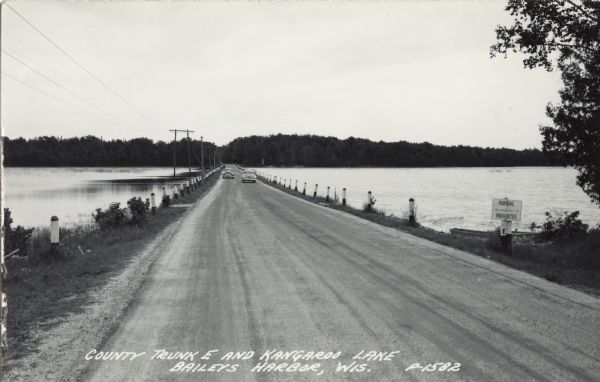 Photographic postcard view of a county road crossing over a lake. Two automobiles are on the road. On the right is a boat at the shoreline behind a "Parking Prohibited" sign. Caption reads: "County Trunk E and Kangaroo Lake, Baileys Harbor, Wis."