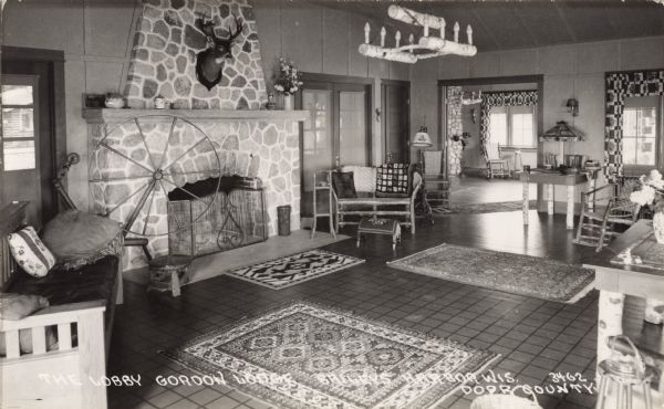 Interior view of the lobby of Gordon Lodge. There is a deer head trophy mounted above the mantle of the stone fireplace, and a large spinning wheel between the fireplace and the door. A candelabra made of logs is hanging from the ceiling in the middle of the room, and there are area rugs on the floor. Caption reads: "The Lobby, Gordon Lodge, Baileys Harbor, Wis. Door County."