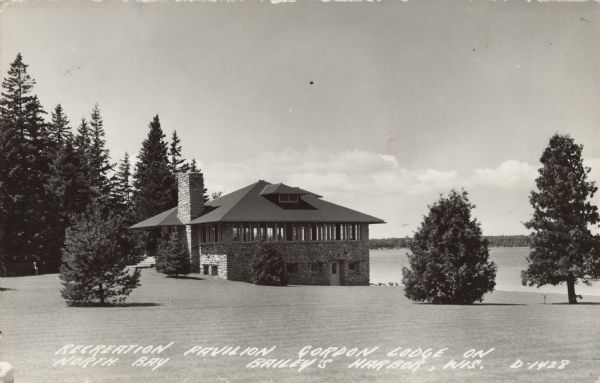 Photographic postcard view across large manicured lawn of the stone building on Lake Michigan. The tree-lined far shoreline is in the background. Caption reads: "Recreation Pavilion, Gordon Lodge on North Bay, Baileys Harbor, Wis."