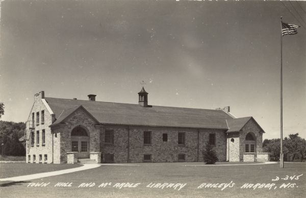 Photographic postcard view of the long stone building that houses the town hall on the right, and the library on the left. On the roof is a small cupola with a weather vane, and there is a fire escape on the back left corner of the building. There is a flag on a flagpole on the front lawn. Caption reads: "Town Hall and McArdle Library,Baileys Harbor,  Wis."