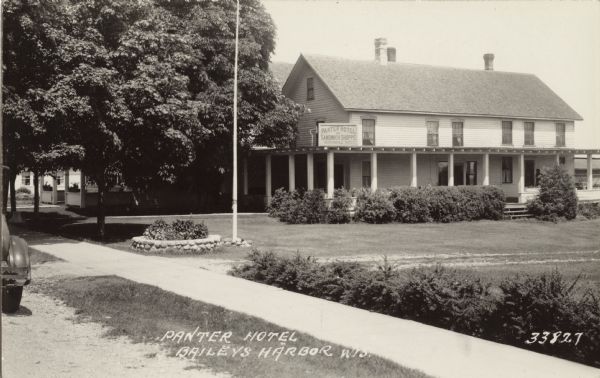 View from road of the Panter Hotel and Sandwich Shoppe, which has a wrap-around porch with bushes planted in front. There are plants in a stone planter in front of the flagpole near the sidewalk Caption reads: "Panter Hotel, Baileys Harbo, Wis."