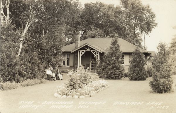 Photographic postcard view of a cottage surrounded by trees and a lawn. A man and woman are sitting in lawn chairs near an oval-shaped stone bordered grouping of plants in the center of the lawn. Caption reads: "Ridge-Birchwood Cottages — Kangaroo Lake, Baileys Harbor, Wis."