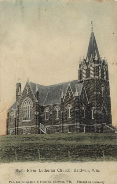 Colorized photographic postcard view of the exterior of the Lutheran Church with arched stained glass windows. There are steps to the arched entrance below the steeple, and also a side entrance. Caption reads: "Rush River Lutheran Church, Baldwin, Wis."