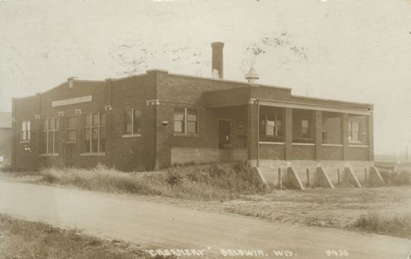 Photographic postcard view across road of the creamery. The entrance is on the left and a loading dock is on the right. Caption reads: "'Creamery,' Baldwin, Wis."