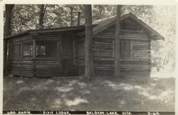 Photographic postcard view of a log cabin in the woods. Caption reads: "Log Cabin, Dixie Lodge, Balsam Lake, Wis."